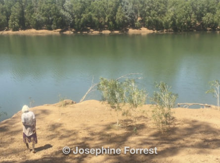 Elderly Aboriginal woman walks down towards the banks of the Martuwarra (Fitzroy River) on a sandy bank with more vegetation visible on the other side of approximately 50m of water.