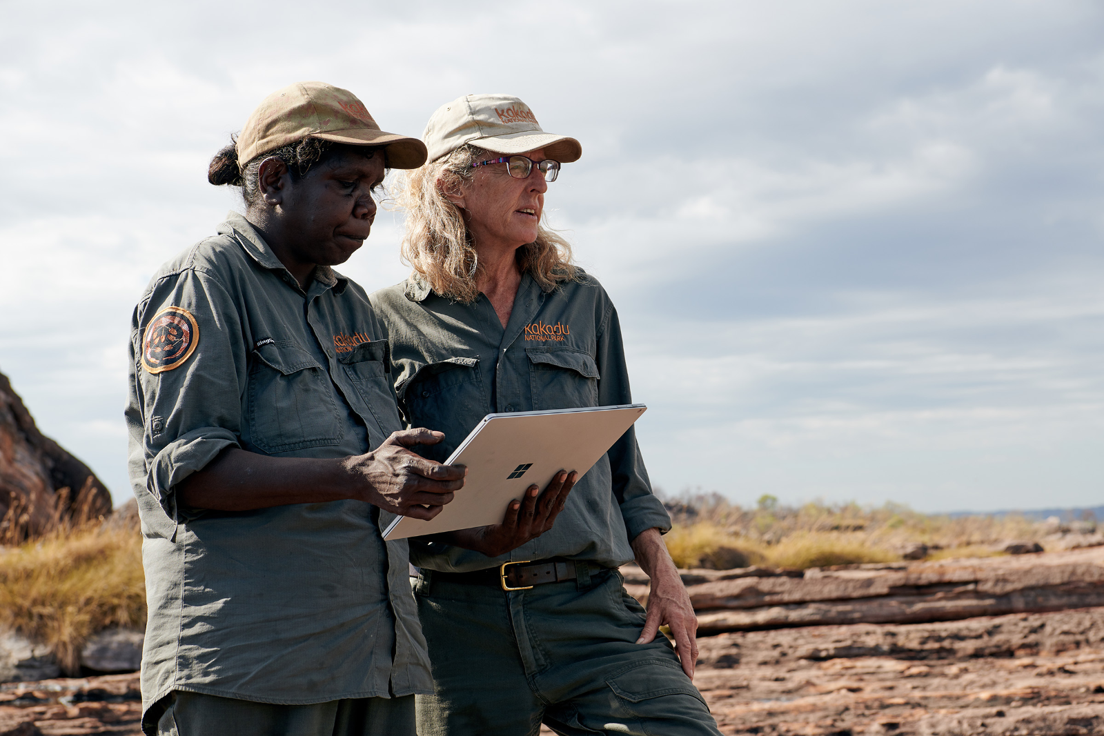 Rangers Serena McCartney and Annie Taylor stand on rocky country looking out over a floodplain and also looking at a Microsoft tablet showing the results of their para grass management on floodplains in Kakadu.