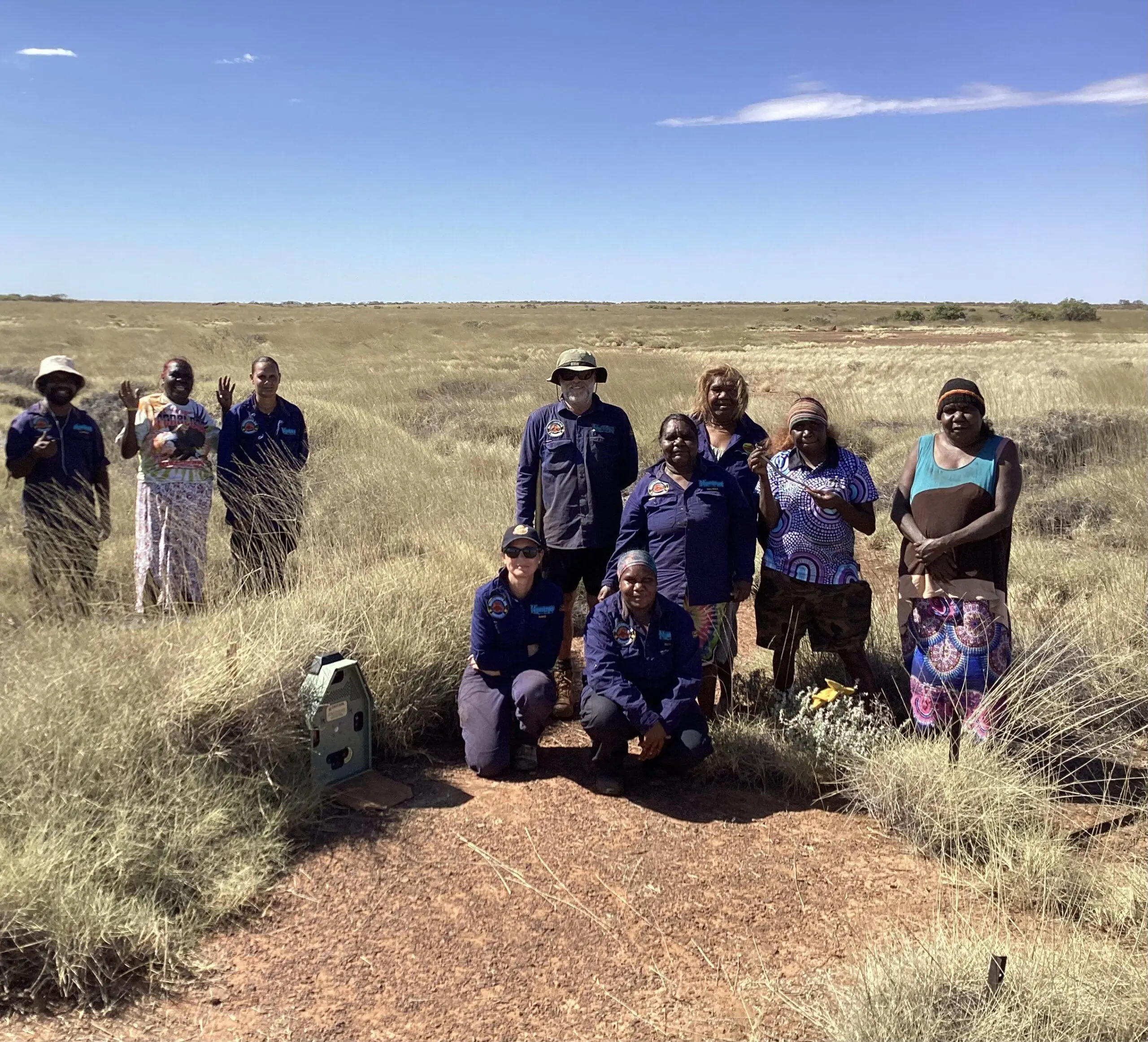 10 people stand in the desert, amongst spinifex which is night parrot habitat. Next to them is a Felixer cat control machine.